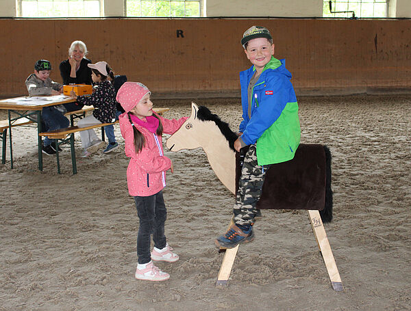 ein Junge sitzt auf einem Holzpferd, vor dem Pferd steht ein Mädchen, im Hintergrund sitzen Personen an einem Tisch