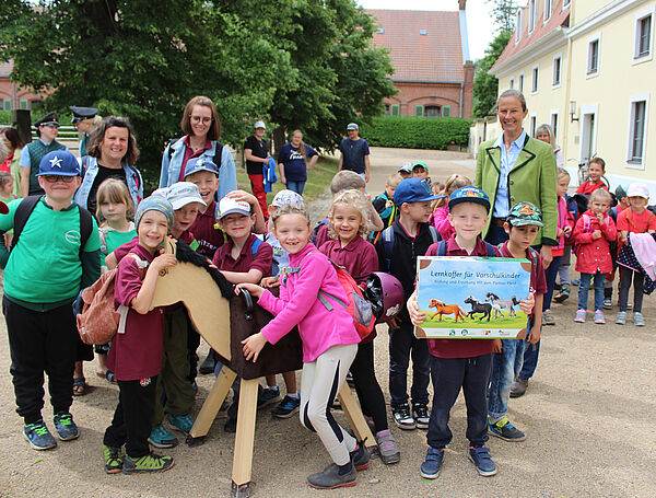 viele fröhliche Kinder gruppieren sich um ein Holzpferd, ein Junge hält einen großen Koffer mit Pferdebildern vor sich