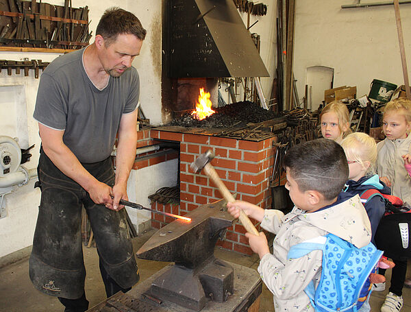 ein Schmied hält ein glühendes Eisen auf einen Amboss, ein Kind schlägt mit einem Hammer auf das Eisen, im Hintergrund sind ein Schmiedefeuer und weitere Kinder