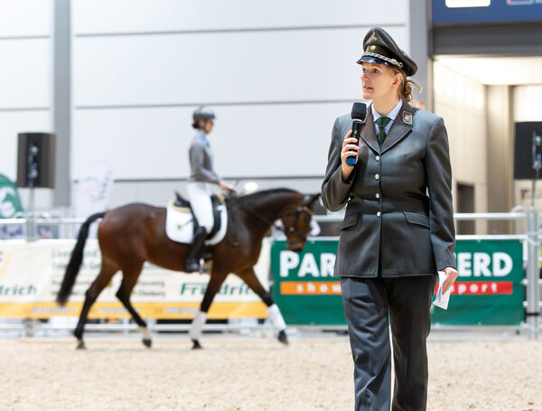 Carolin Dins spricht ins Mikro, im Hintergrund reitet eine Auszubildende